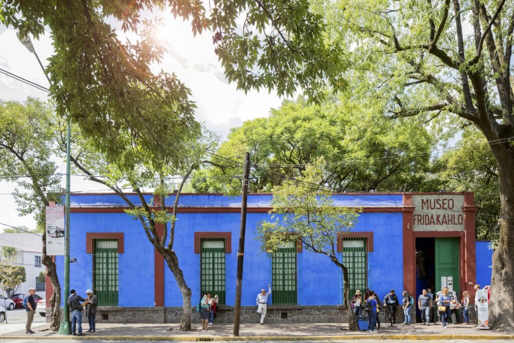 People line up in front of a one-story building painted blue on a city street shaded by trees.