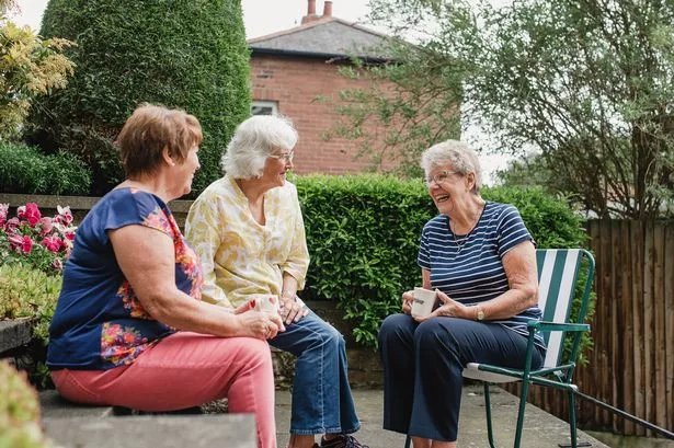 Three senior women are sitting in the garden with a cup of tea while they chat and laugh together.
