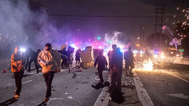 Demonstrators in jumpers run away from police as fireworks go off, siren lights illuminate the night