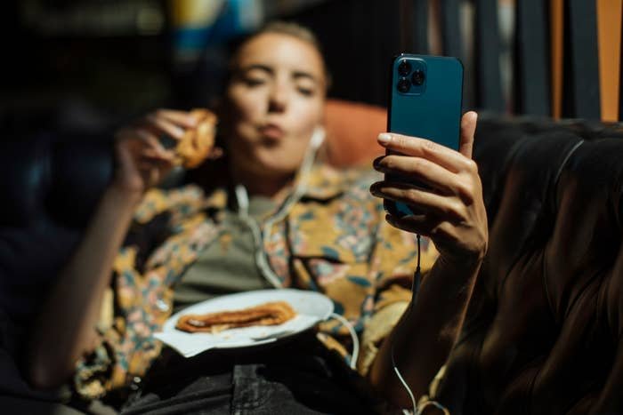 Person lounging on a couch, eating a pastry and taking a selfie with a smartphone, earphones plugged in