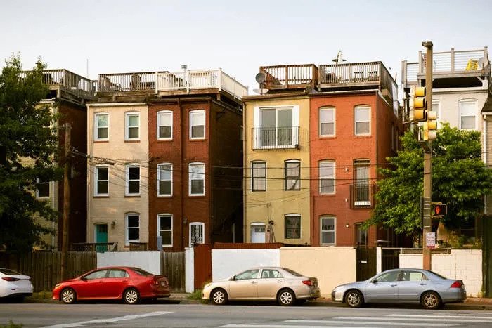 Three brick row houses with rooftop decks are lined up on a city street, with four parked cars in front. Traffic light on the corner