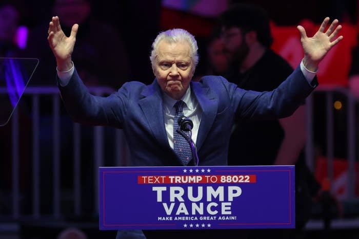 Man on stage raising arms, wearing a suit, stands behind a podium with "Trump Vance" and a phone number, during a public speaking event