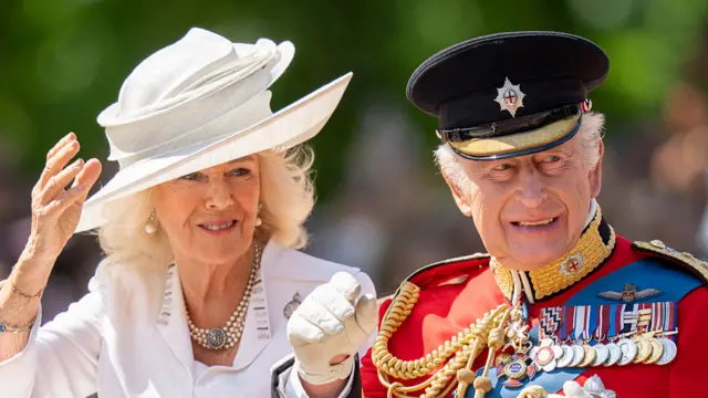 Charles wears red uniform and a black hat alongside Camilla who wears a white blazer and hat.