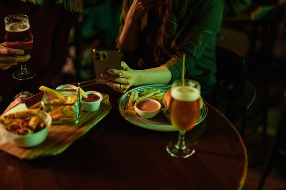 Young women enjoying drinks and food while looking at a smart phone in a lively pub, sharing laughter and moments of friendship