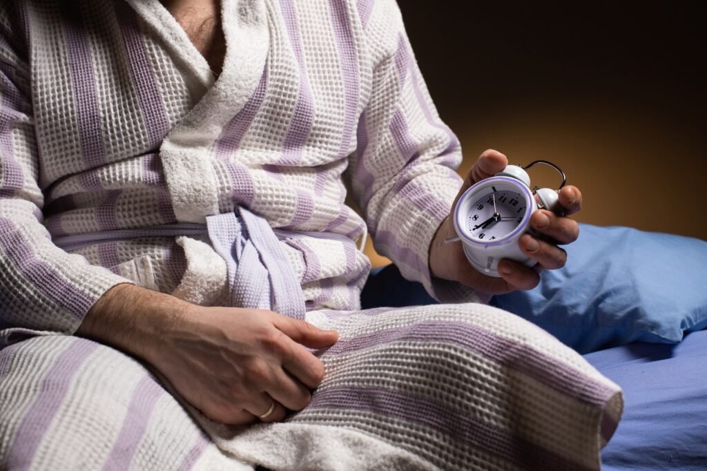 A man in a bathrobe holds an alarm clock in his hand