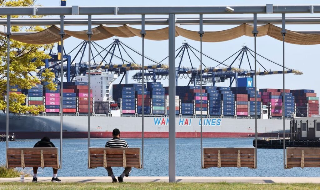 People sit in a park near shipping containers stacked on a container ship at the Port of Los Angeles on June 25, 2025.
