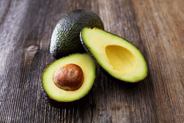 Close-up of two avocados on a wood surface; one is whole, the other is sliced in half and laid open in front of it.