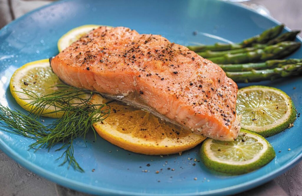 A serving of salmon sprinkled with seasonings, plated on slices of lemon and lime with spears of asparagus alongside it.
