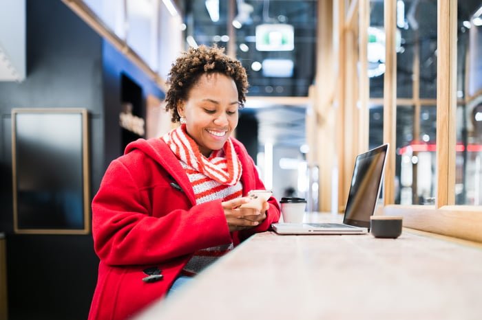 A person smiles while looking at a cell phone and sitting at a table in a public setting with a hot beverage and a laptop computer.