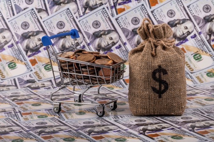 A shopping cart full of coins next to a bag of money on top of hundred-dollar bills.