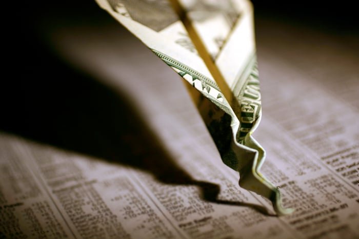 A twenty dollar bill paper airplane that's crashed and crumpled into a financial newspaper.