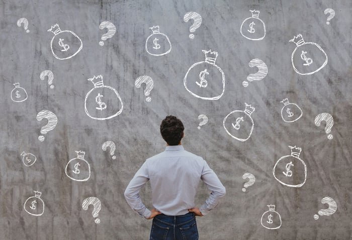 man before a wall with question marks and money bags.