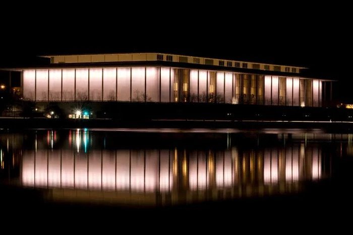 An illuminated large building at night is reflected in a calm body of water, showcasing its architectural design