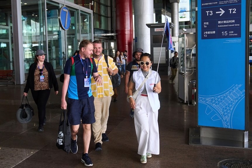 woman in white shirt, trousers, oversized sunglasses carrying small blue flag exits airport with two tourists with backpacks