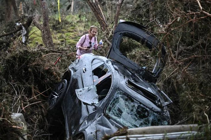 Person in rugged terrain stands beside an overturned car with broken windows. Dense branches surround the scene, suggesting an accident