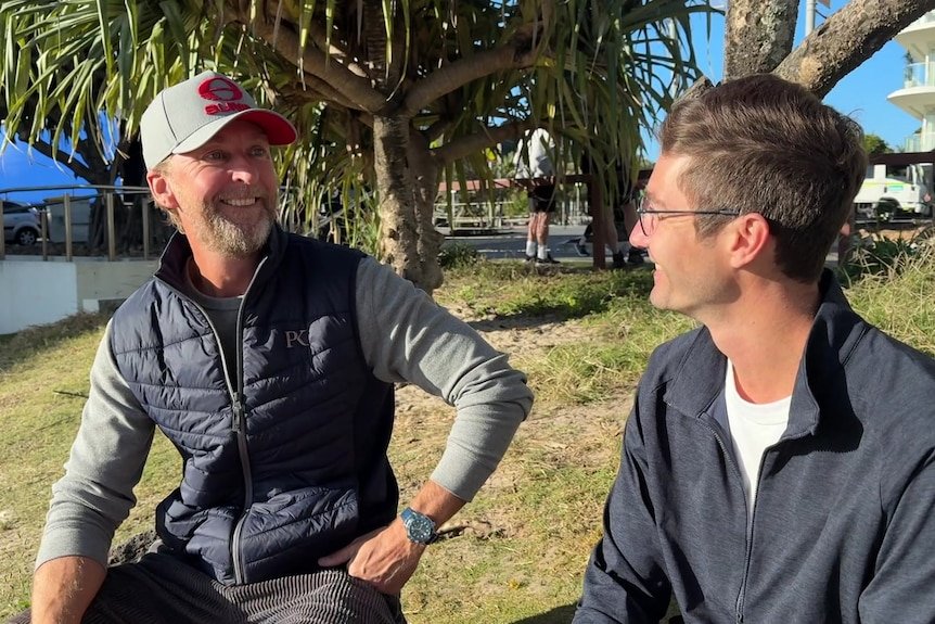Two men smile at each other while sitting outdoors on a sunny day.