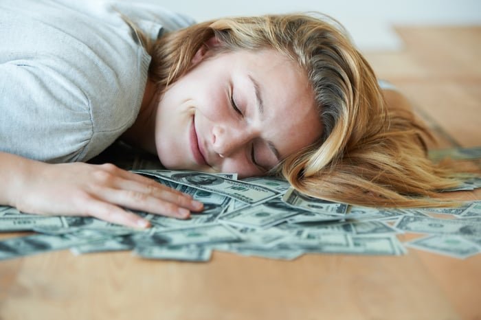 A smiling person sleeps on a heap of dollar bills.