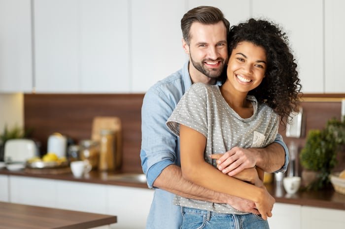 A couple is smiling in a kitchen, one holding the other.