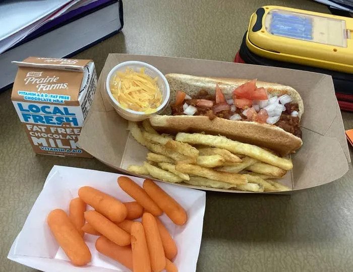 Computer screen showing a Reddit post with a photo of a school lunch: hot dog, fries, carrots, fruit cup, and milk