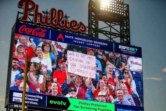 A crowd at a Phillies game is shown on the big screen holding a humorous sign about not cheating on their significant others