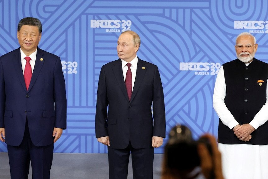 Three men stand side-by-side in suits in front of a blue board