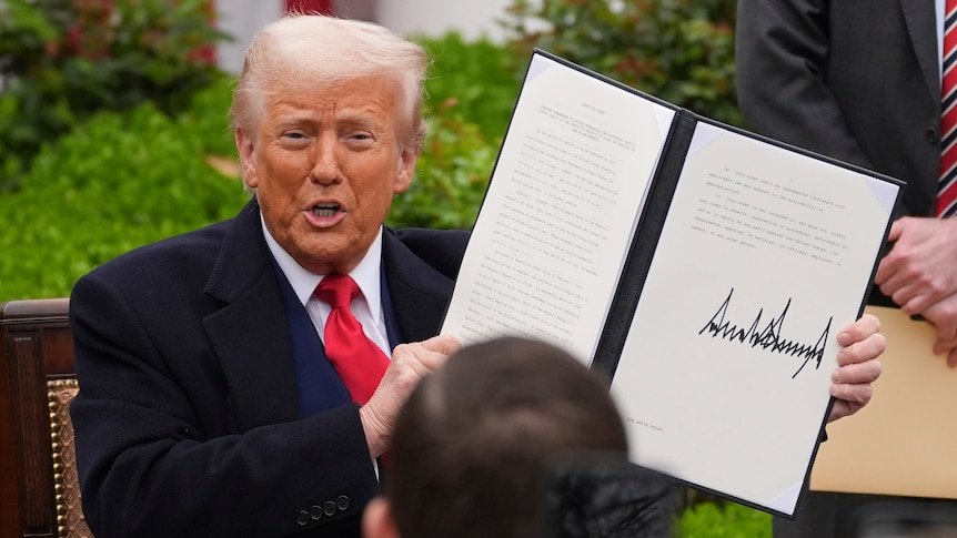 Donald Trump wears a blue suit, white shirt and red tie holding a booklet showing off his signature to the camera
