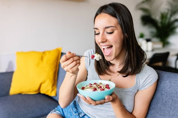 Beautiful young woman having breakfast with bowl of yogurt, cereals and fruits sitting on sofa at home.
