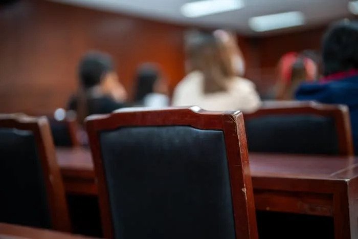 People seated in a courtroom setting, viewed from behind, focusing on the back of a chair
