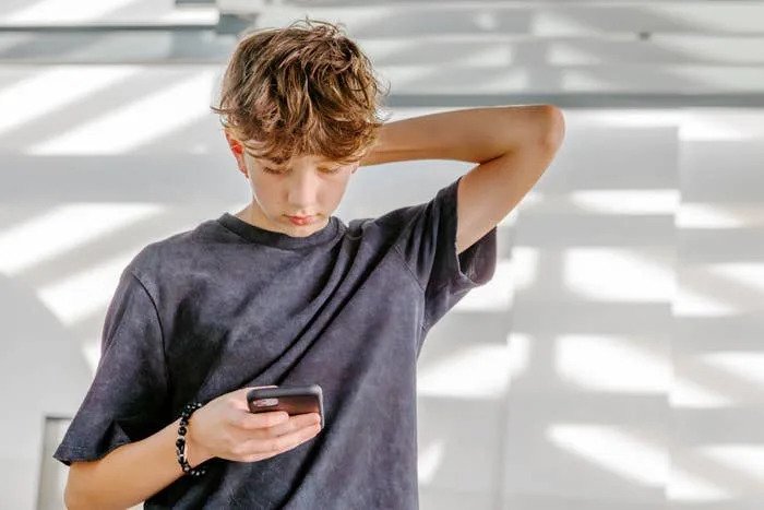 Teen looking down at a phone, hand behind head, standing in a room with patterned shadows on the wall. Casual attire