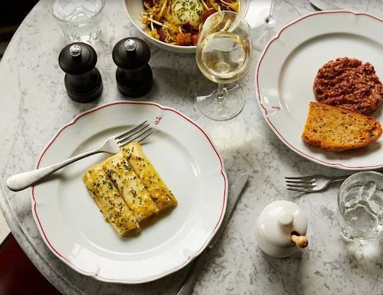 Table with three dishes including roasted asparagus and beef tartare, a glass of white wine, and two salt and pepper shakers.