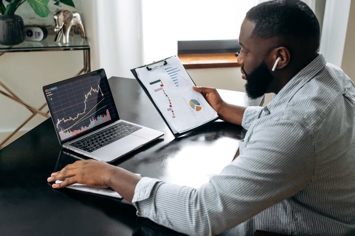 An investor reviews a clipboard with a chart along with a laptop showing stock price information.