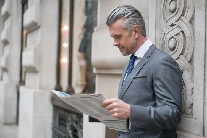 A business person in a gray suit reads a newspaper while leaning against a stone wall.
