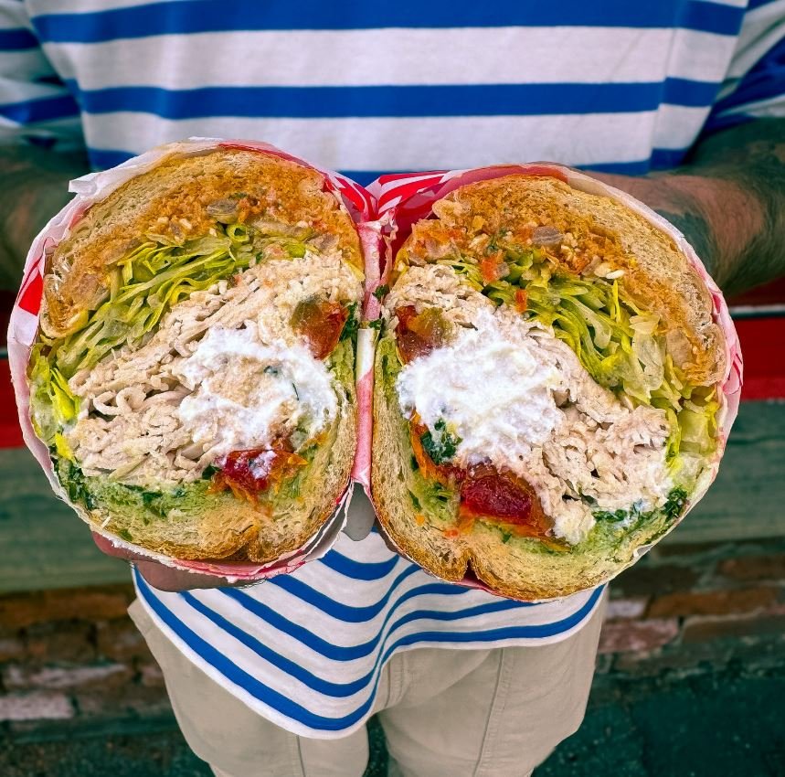 Large sub sandwich, cut in half, filled with shredded meat, lettuce, sauce, and red relish, held by a person.