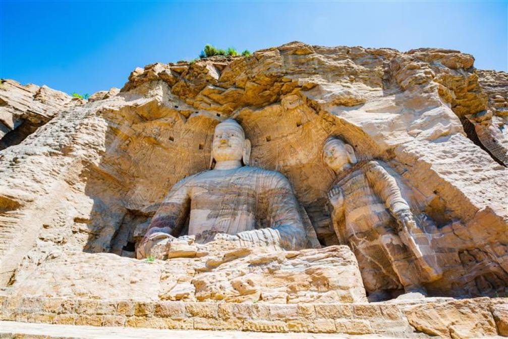 Two large stone Buddha statues carved into a cliff face, with intricate details and serene expressions, under a clear blue sky.