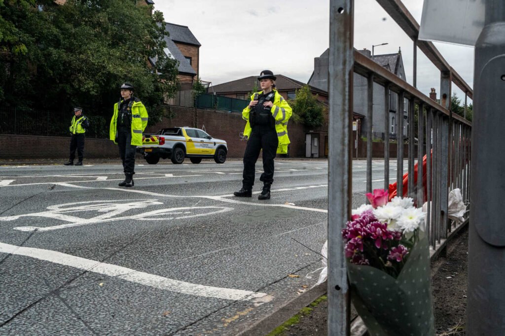 Three police officers stand on a road with a fence with flowers leaned against it in the foreground.