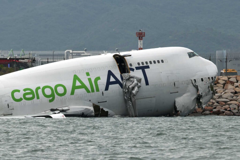 A damaged plane rests against a wall of rocks next to a body of water.