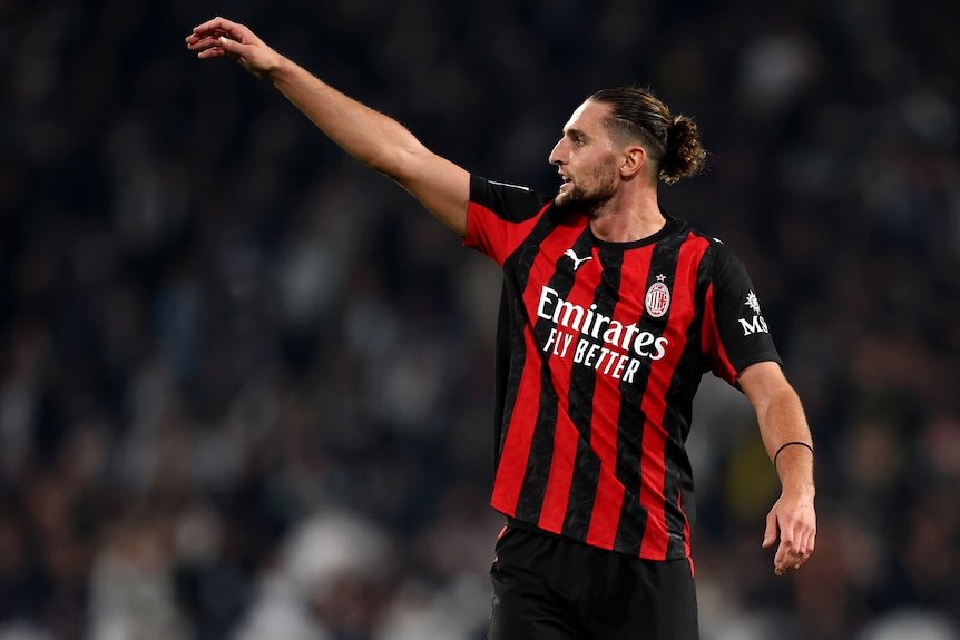 A footballer waves his hand in the air while on the field, wearing a red and black striped jersey.