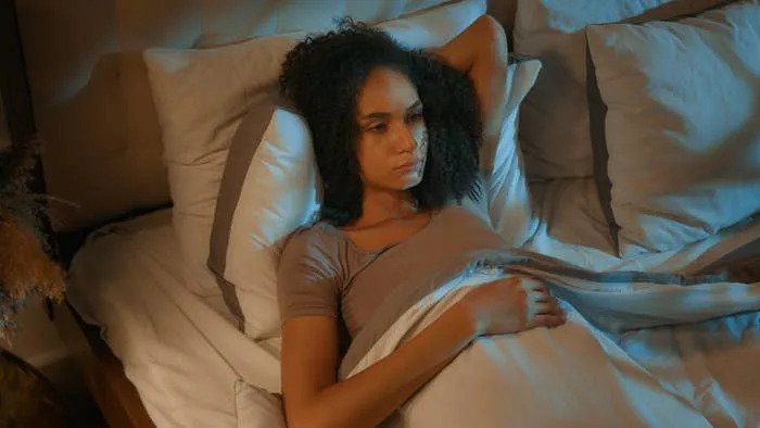 Person lying in bed, looking pensive and lost in thought, surrounded by pillows and soft bedding