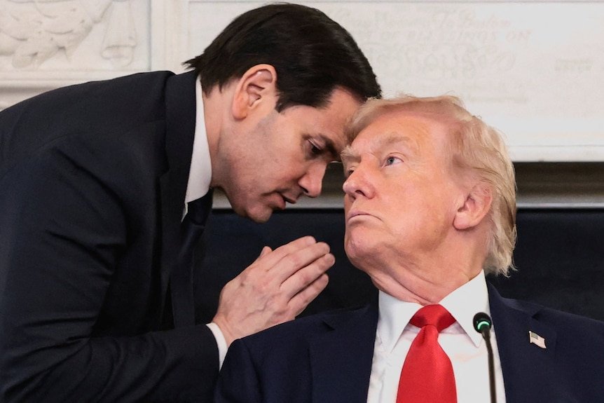 A dark-haired man leans in close to speak to Donald Trump. Both wear dark suits.