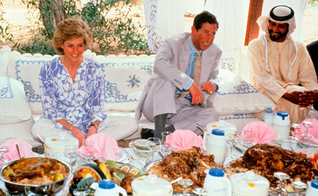 Princess Diana sitting on a cushion next to Prince Charles in front of a big table of food