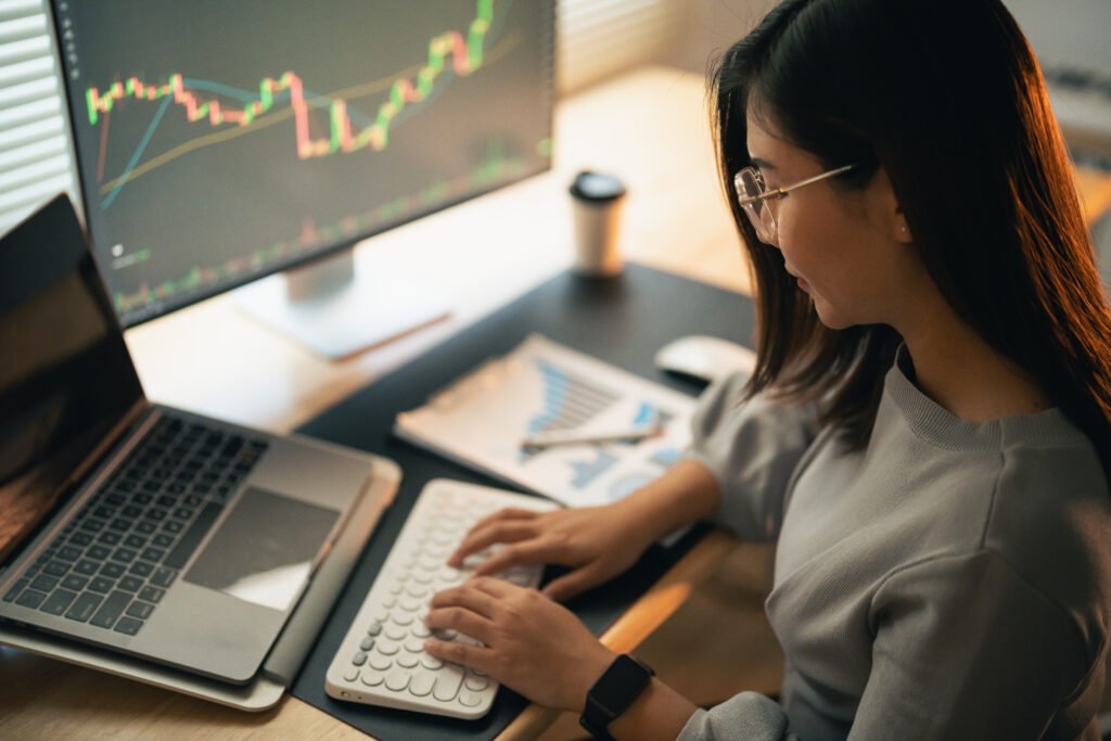 A person sits at a desk using their laptop and looks at a stock chart on a second monitor.